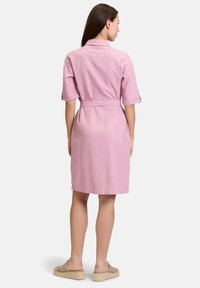 Różowa lniana sukienka z krótkimi rękawami i paskiem w talii. Widok z tyłu ukazuje prosty krój i długość do kolan. Założona z beżowymi espadrylami.