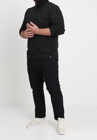 Pull-over noir en maille torsadée avec col zippé, associé à un pantalon noir et des baskets blanches. La maille présente un motif texturé.