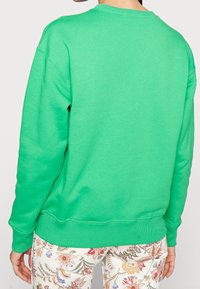 Sudadera verde hecha de tela suave con cuello redondo y puños acanalados. Llevada con pantalones estampados de flores que muestran colores brillantes.