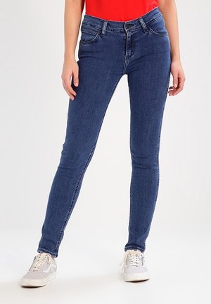 Kvinde iført blå skinny jeans, hvide sneakers og en rød top, stående med fødderne let adskilt på en ensfarvet baggrund.