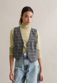 Szara kamizelka w kratę z żółtymi paskami, z wciętym ko�łnierzem i guzikami z przodu, noszona na jasnozielonym golfie i wyblakłych niebieskich dżinsach.