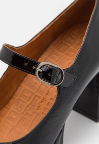 Chaussure Mary Jane en cuir verni noir avec une boucle argentée arrondie et une semelle intérieure rembourrée couleur fauve embossée d'un motif géométrique.
