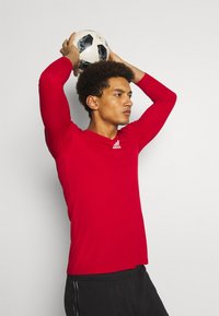 Rode sportshirt met lange mouwen en ronde hals, met het witte Adidas-logo. Model houdt een zwart-witte voetbals boven zijn hoofd.