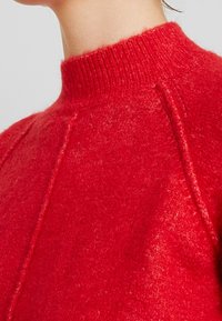 Roter Strickpullover mit hohem, geripptem Kragen, weicher Textur und Raglanärmeln, der einen tiefen Farbton und subtile Variationen im Stoff zeigt.