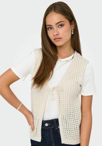 Gilet senza maniche lavorato a maglia di colore crema con apertura davanti, motivo testurizzato e chiusura con fiocco, indossato sopra una camicia bianca e jeans scuri.