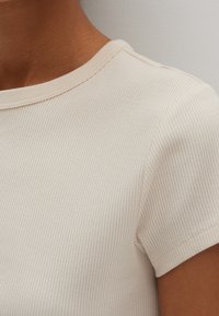 Beige ribbad t-shirt med rundad halsringning och korta ärmar, som visar upp ett texturerat tyg och subtila sömdetaljer längs sömmarna.