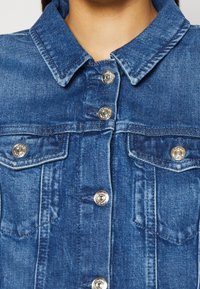 Giacca di jeans blu con colletto classico, tasche frontali e bottoni in argento. Il tessuto ha un aspetto leggermente strutturato.