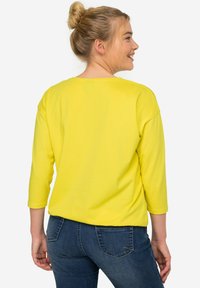 Gelbes, locker sitzendes Langarmshirt mit weitem Halsausschnitt und bequemen Schultern, kombiniert mit blauen Jeans, von hinten betrachtet.
