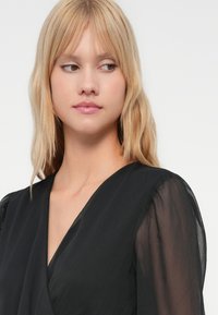 Blondi nainen, jolla on suorat hiukset ja musta läpinäkyvähiha, katsoo vasemmalle yksinkertaista vaaleata taustaa vasten.