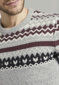 Homme portant un pull tricoté à motifs gris, bordeaux, noir et blanc avec des dessins en zigzag et géométriques sur fond gris clair.