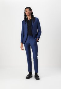 Costume bleu avec un blazer ajusté et un pantalon slim, associé à une chemise noire. Tissu lisse avec un éclat subtil et des détails traditionnels de boutons.