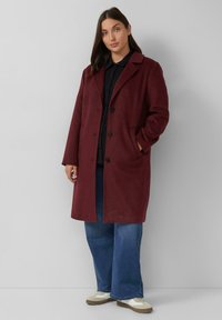 Bordeaux wollen jas met een reverskraag, enkele knopen aan de voorkant en twee zijzakken, gedragen over een donker shirt en wijde spijkerbroek.