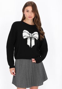 Pulover negru cu grafică de fundiță albă, tricotat texturat și manșete cu nervuri, asociat cu o fustă gri plisată. Combinație simplă și casuală de outfit.