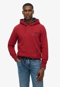 Felpa rossa con cappuccio, tasca frontale, cordini e dettaglio del logo. Abbinata a jeans blu e un orologio, presenta un look informale.