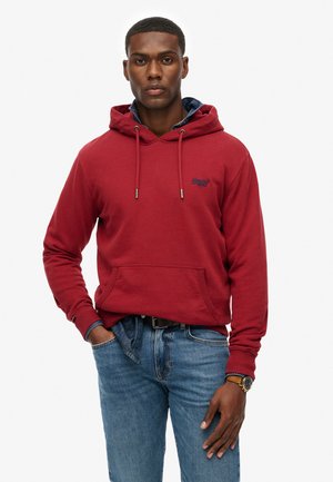 Red hooded sweatshirt with front pocket, drawstrings, and logo detail. Paired with blue jeans and a watch, presenting a casual fit.
