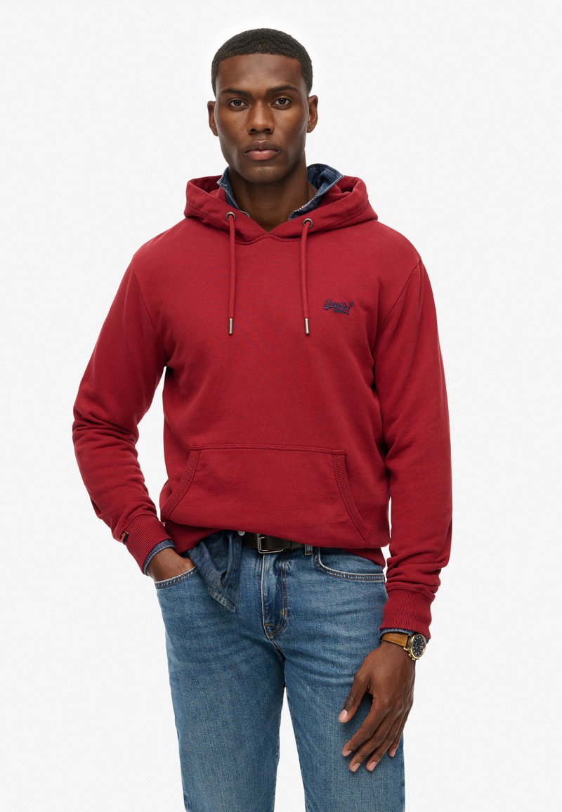 Felpa rossa con cappuccio, tasca frontale, cordini e dettaglio del logo. Abbinata a jeans blu e un orologio, presenta un look informale.
