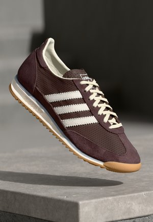 Mørkebrun sneakers med hvide striber og snørebånd, lys beige sål med gummimønstret slidbane, svævende over en grå betongoverflade.