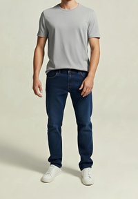 Mann trägt ein hellgraues kurzärmliges T-Shirt, dunkelblaue Jeans und weiße Turnschuhe, steht vor einem schlichten beigefarbenen Hintergrund.