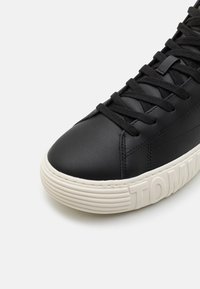 Svarta lädersneakers med svarta snören och tjock vit gummisula med delvis präglat varumärkesnamn, visat mot en vit bakgrund.