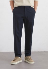 Marineblaue Chinos aus Baumwolle, mit schmaler Passform und seitlichen Taschen, kombiniert mit hellbraunen Wildledersneakern auf einem hellgrauen Boden.