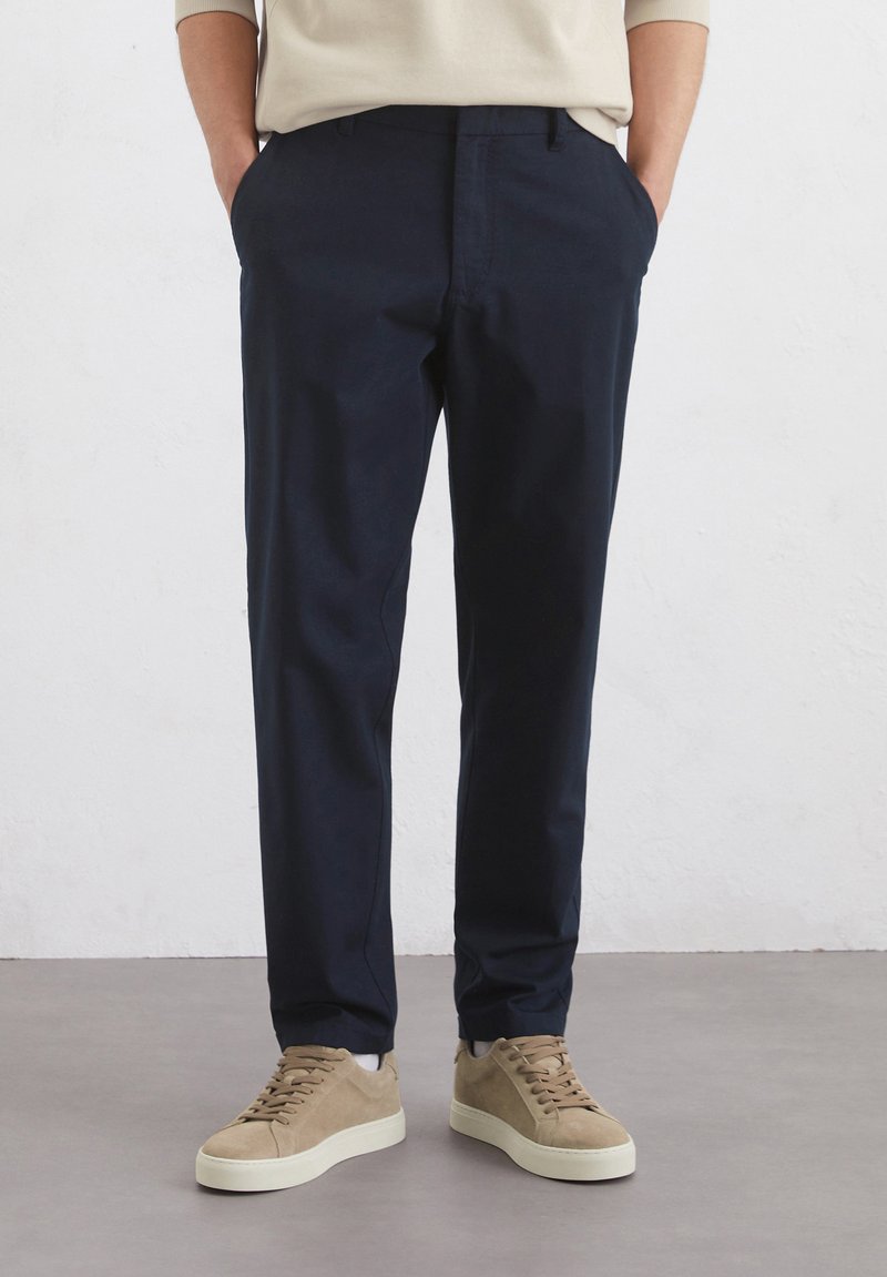 Marineblaue Chinos aus Baumwolle, mit schmaler Passform und seitlichen Taschen, kombiniert mit hellbraunen Wildledersneakern auf einem hellgrauen Boden.