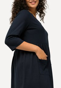 Granatowa sukienka z miękkiego materiału, z dekoltem w serek, rękawami do trzech czwartych, marszczonym pasem i kieszenią po boku.