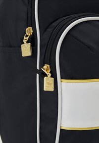 Czarny nylonowy plecak z złotymi suwaki i białymi akcentami, z fakturowaną powierzchnią i wyraźnym detalem logo.