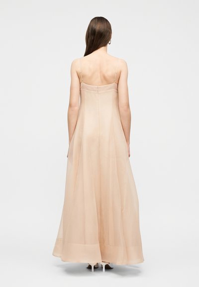 Γυναίκα με μακριά καστανά μαλλιά, φορώντας ένα μπεζ φόρεμα με λεπτές τιράντες που φτάνει μέχρι το πάτωμα και λευκά παπούτσια με τακούνι, στέκεται μπροστά σε ένα απλό ανοιχτόχρωμο φόντο.