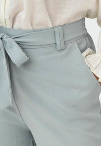Pantalon gris clair en tissu texturé, avec une taille ceinturée et un nœud. Comprend des poches latérales et une coupe décontractée.