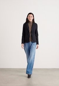 Schwarze Jacke mit zwei Fronttaschen, kombiniert mit einem gemusterten Rollkragenpullover und lockeren blauen Jeans. Tragen mit schwarzen, hohen Stiefeletten.