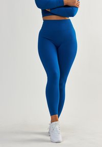 Leggings bleus taille haute en tissu extensible, texture lisse, design long, associés à des baskets blanches.