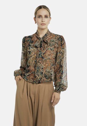 Frau trägt eine durchsichtige Bluse mit Paisley-Muster, einem Schleifenkragen und beigen, hoch taillierten, plissierten Hosen, posiert mit einer Hand in der Tasche.