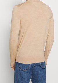 Pull beige en maille avec manches longues et poignets côtelés, doté d'un col rond et d'une texture lisse, associé à un jean en denim bleu.