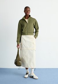 Olivengrønn zip-up jakke, beige midi skjørt med lommer, hvite sneakers med svarte accenter, og en grønn skulderveske. Jevn tekstur gjennomgående.
