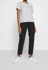 T-shirt grigia in cotone, vestibilità rilassata, abbinata a jeans neri a gamba dritta e sneaker bianche, su sfondo bianco semplice.