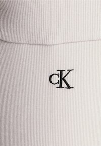 Vaaleanharmaa ribattua kangasta, jossa on musta brodeerattu "ck"-logo. Materiaali on sileä ja muotoon sulautuva.