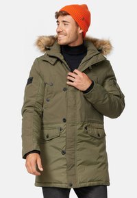 Parka verde oliva con capucha forrada de piel, que cuenta con múltiples bolsillos frontales, cierres de botones y acentos en negro. Diseño cálido e insulated.