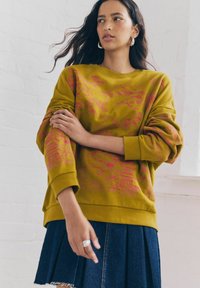 Sweatshirt vert olive avec un motif floral rose, coupe décontractée, manches larges et poignet côtelé. Associé à une jupe plissée en denim.