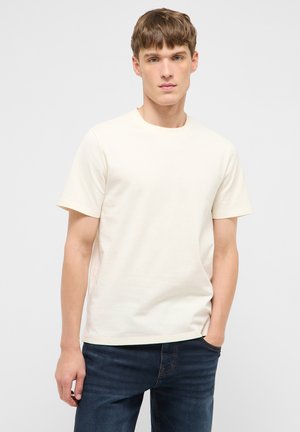 Junger Mann mit kurzen braunen Haaren, der ein schlichtes cremefarbenes T-Shirt mit kurzen Ärmeln und dunkelblaue Jeans trägt, steht mit einer Hand in der Tasche.