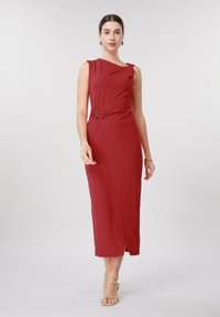 Γυναίκα που φοράει αμάνικο εφαρμοστό κόκκινο μίντι φόρεμα με λεπτομέρεια συγκέντρωσης στο πλάι και μπεζ πέδιλα με λουράκια, όρθια μπροστά σε απλό φωτεινό φόντο.