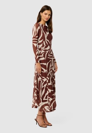 Femme portant une robe midi à manches longues, marron et crème, à motif abstrait, avec des talons à bride cheville, se tenant devant un fond gris clair.