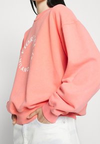 Koraalroze sweatshirt met een ronde halslijn, oversized pasvorm, lange mouwen en witte logo print op de voorkant. Zacht katoenmateriaal.