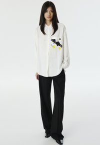 Chemise blanche à boutons, en tissu texturé, ornée de broderies florales noires et jaunes sur la poche poitrine gauche, associée à un pantalon noir.