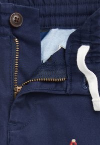 Närbild av marinblå byxor med delvis öppen mässingsdragkedja, brun knapp, vit dragsko och ljusblå innertyg synligt.