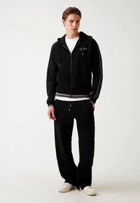 Hanorac negru cu fermoar, cu dungi albe pe manșete și tiv, purtat peste un tricou alb și asortat cu pantaloni sport negri și încălțăminte deschisă la culoare.