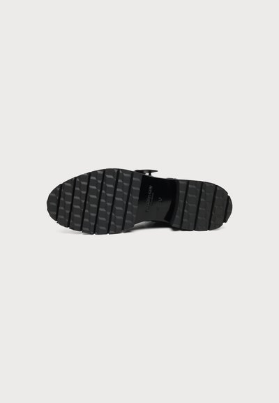 Semelle de chaussure noire avec des rainures horizontales profondes et larges et une partie centrale brillante, présentée sur un fond clair uni.