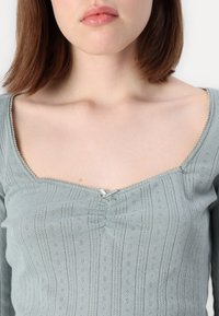 Vaaleanvihreä lyhyt toppi, jossa on teksturoitu kangas, neliömäinen kaula-aukko, koristeompeleet ja pieni nauhakiinnitys keskellä.