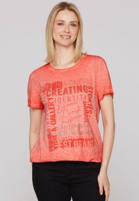 Camiseta de manga corta en color coral con tipografía mixta y elementos gráficos en un tono más claro. Tejido suave y texturizado. Corte casual.