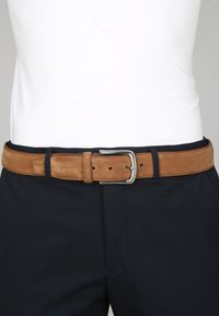Bruine leren riem met zilveren gesp, gedragen om de taille over zwarte broek en een ingetrokken wit shirt.
