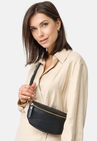 Zwarte leren crossbodytas met een gestructureerd oppervlak en gouden rits, vastgehouden door een persoon in een lichtbeige shirt.